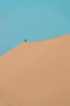 A lone person on the top of a sand dune against a blue sky.