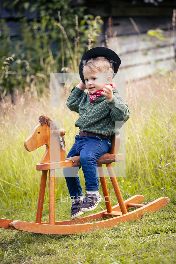 Little Cute Boy in Cowboy Costume on Rocking Horse in the Green Summer Forest