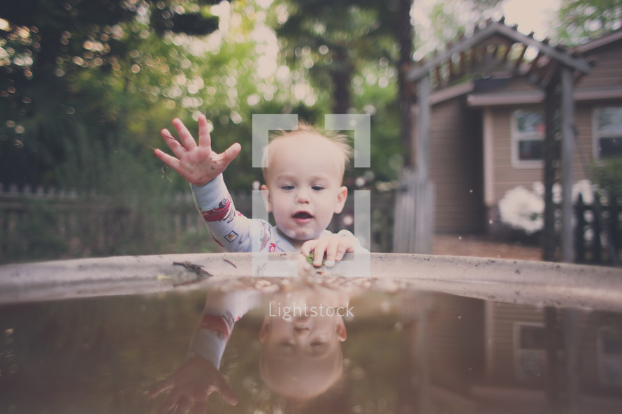 A baby boy plays in the water of a birdbath.