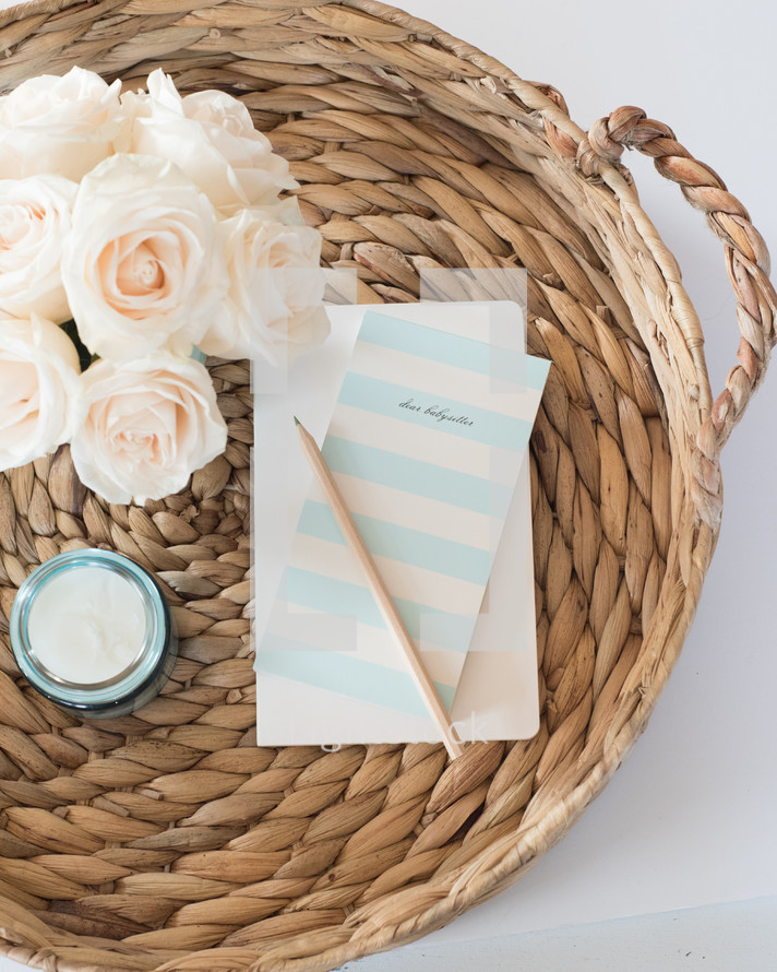 A pencil and notecard, candle, and white flowers on a rattan basket.