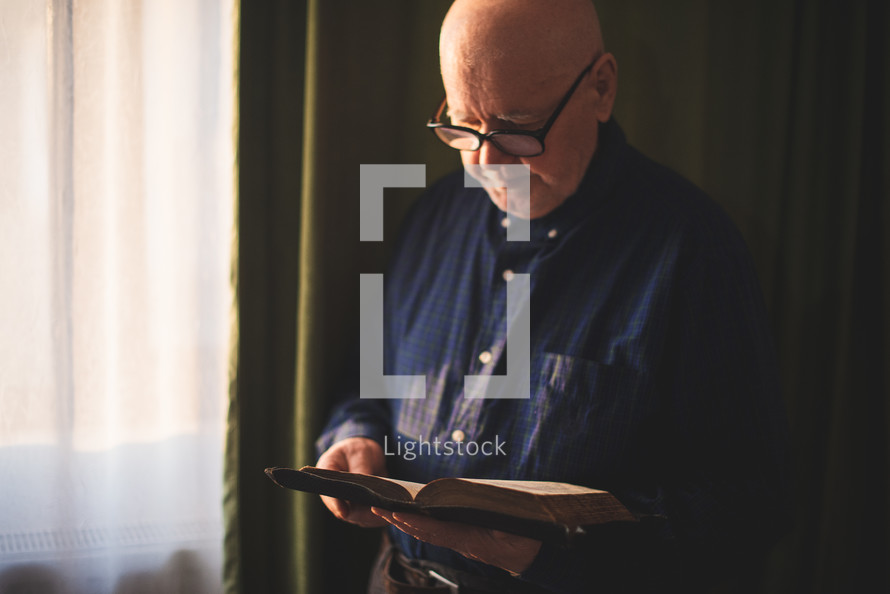 an elderly man reading a Bible standing in a window 