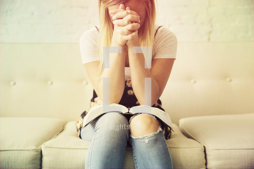 woman sitting on a couch in prayer with an open Bible in her lap.