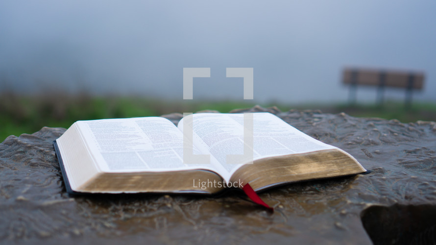 Close up shot of a open bible on a podium outside on a cloudy day with bench in the background.