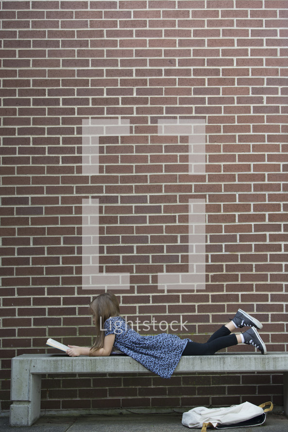 a child sitting on a bench reading a book while waiting for school to start 