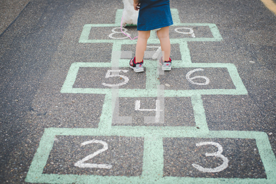 A young girl standing on a hopscotch board with a stuffed animal. 