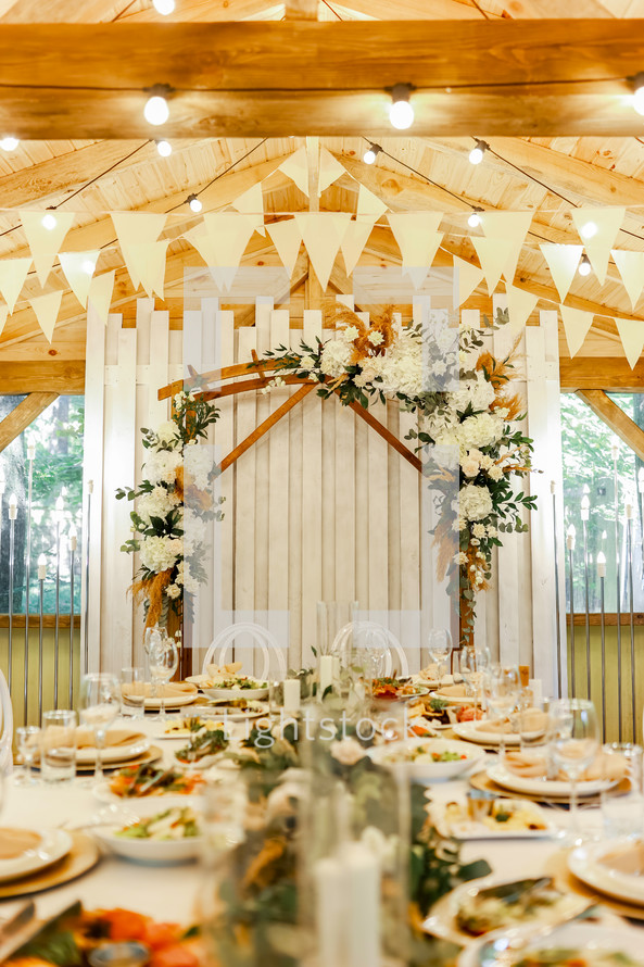 Festive setting table with meals for wedding, wooden arch, stands decorated with composition of white flowers and greenery in the rustic banquet hall