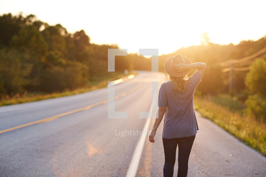 a woman in a hat walking down a country road 