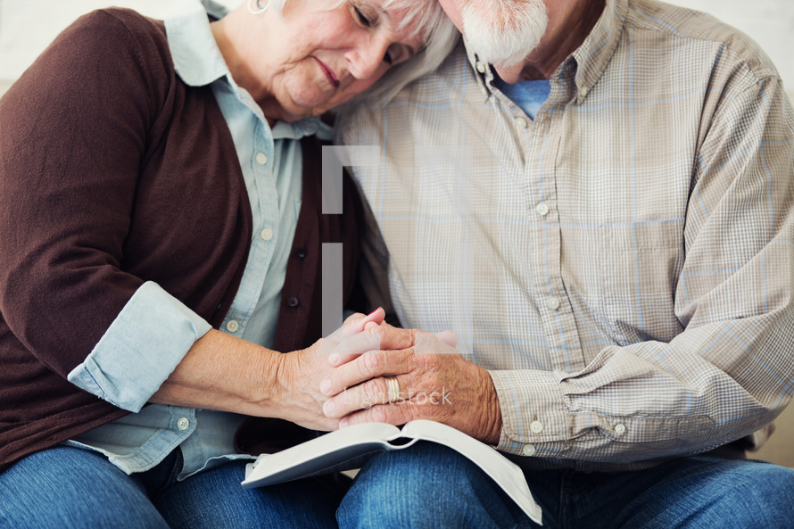 elderly couple holding hands and praying over the pages of a Bible 