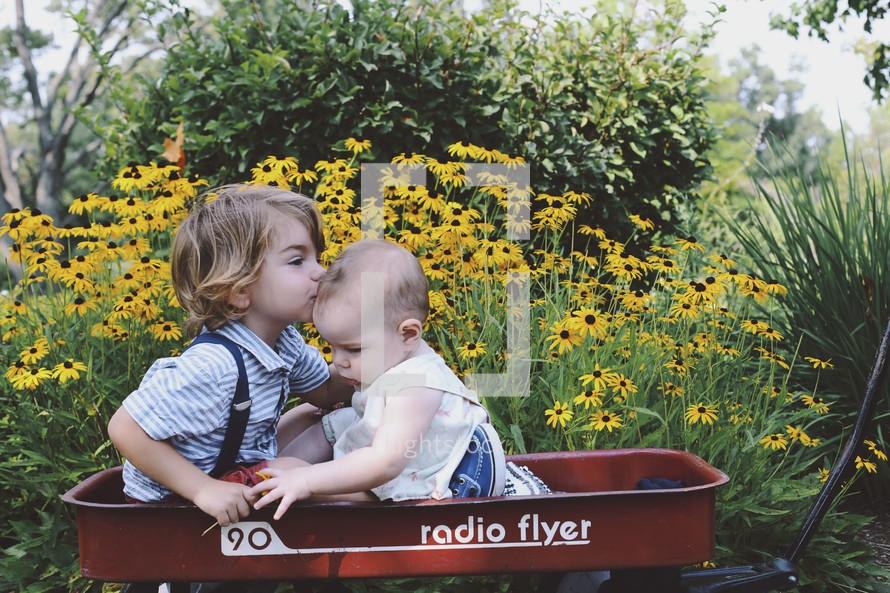 brother and sister in a red wagon near yellow flowers 