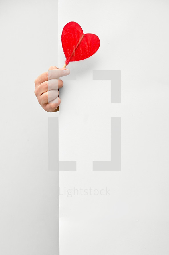 Male hand holding and offering Heart Shaped Lollipop behind white wall background