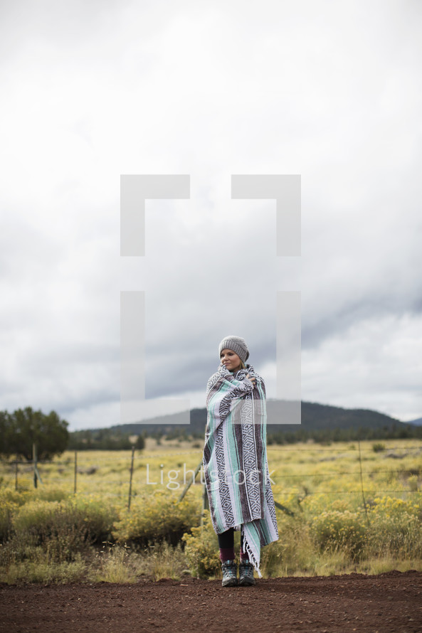 A woman wrapped in a blanket stands on a gravel road near a field.