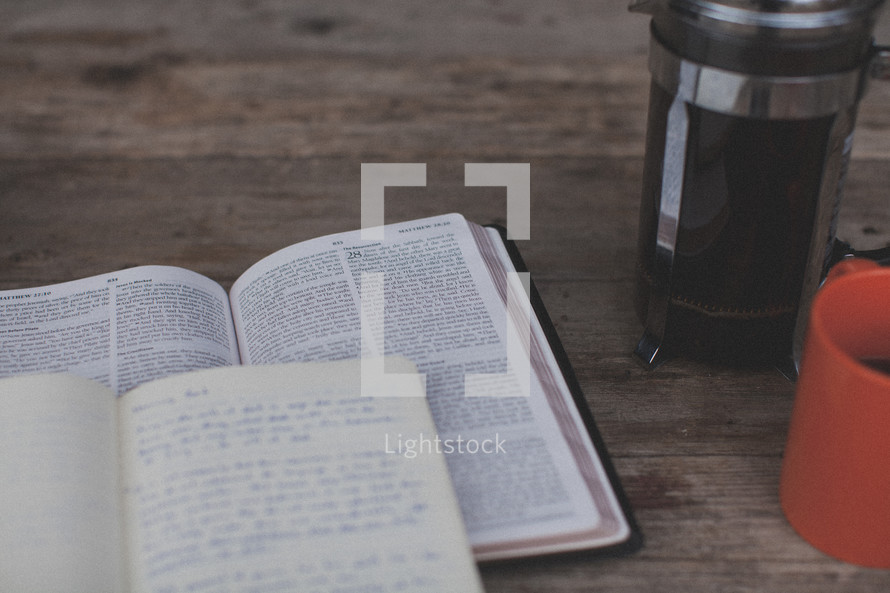 An open Bible and notebook next to an orange coffee mug and french press
