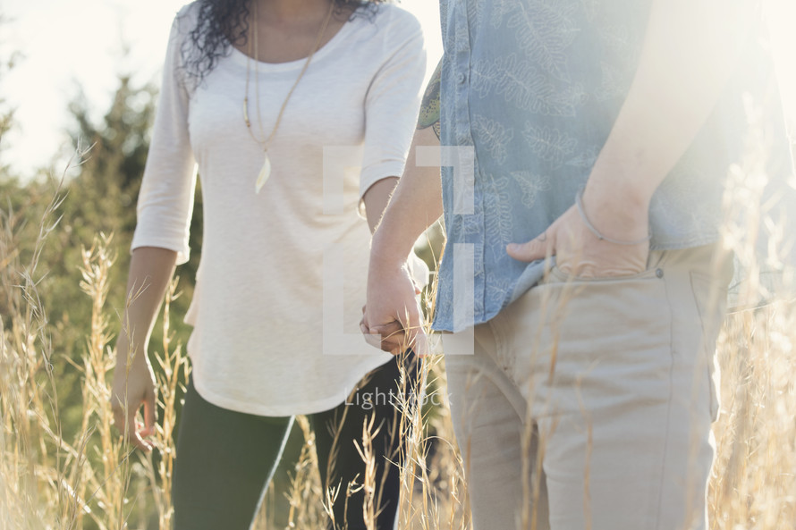 a couple walking holding hands through tall grasses 