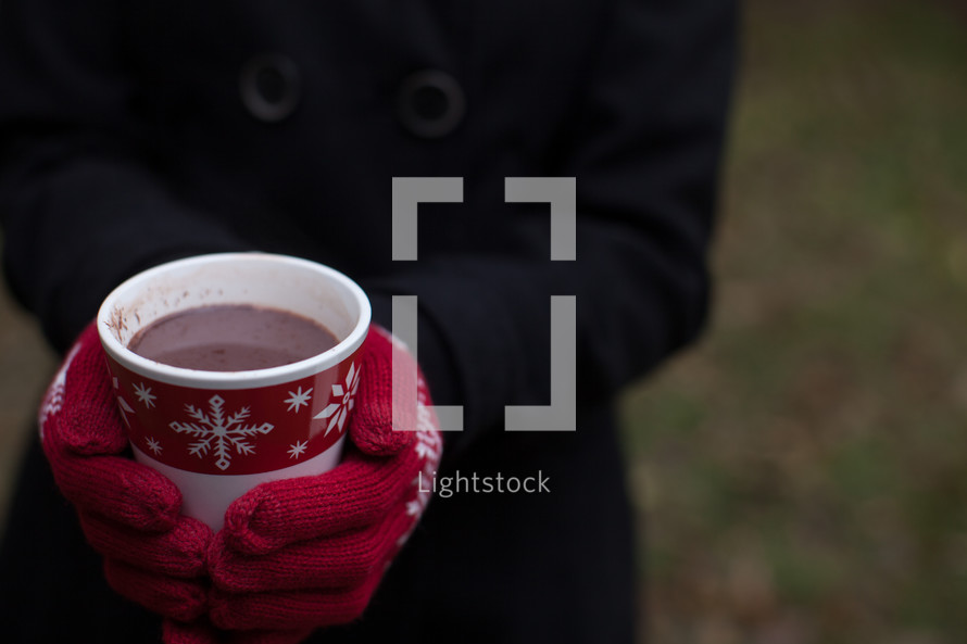 A cup of hot chocolate in a Christmas cup being held by a woman wearing red mittens.