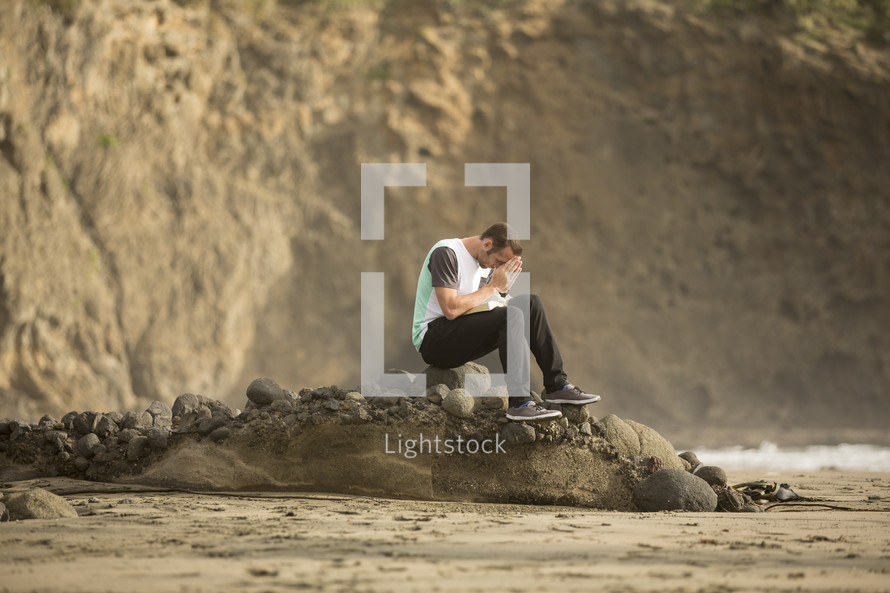 man sitting on a rock with head bowed in prayer on a beach 