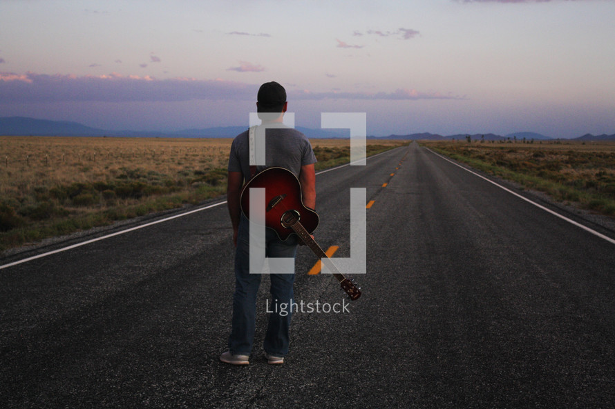man standing in the middle of a road with a guitar at sunset 