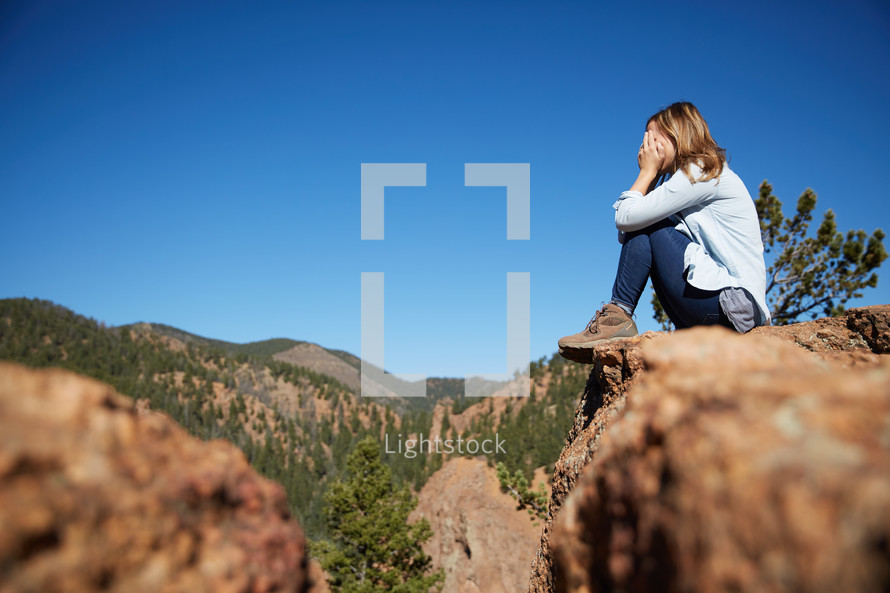 a woman covering her face sitting on the edge of a mountain 