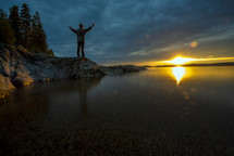 a man with raised hands standing on a rocky shore 