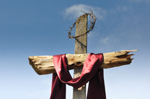 Wooden cross with crown of thorns and red satin fabric against a blue sky.