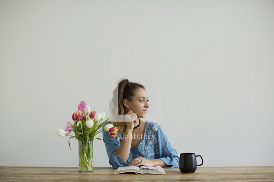 a woman sitting at a desk with a mug of coffee and a vase of tulips reading a Bible 