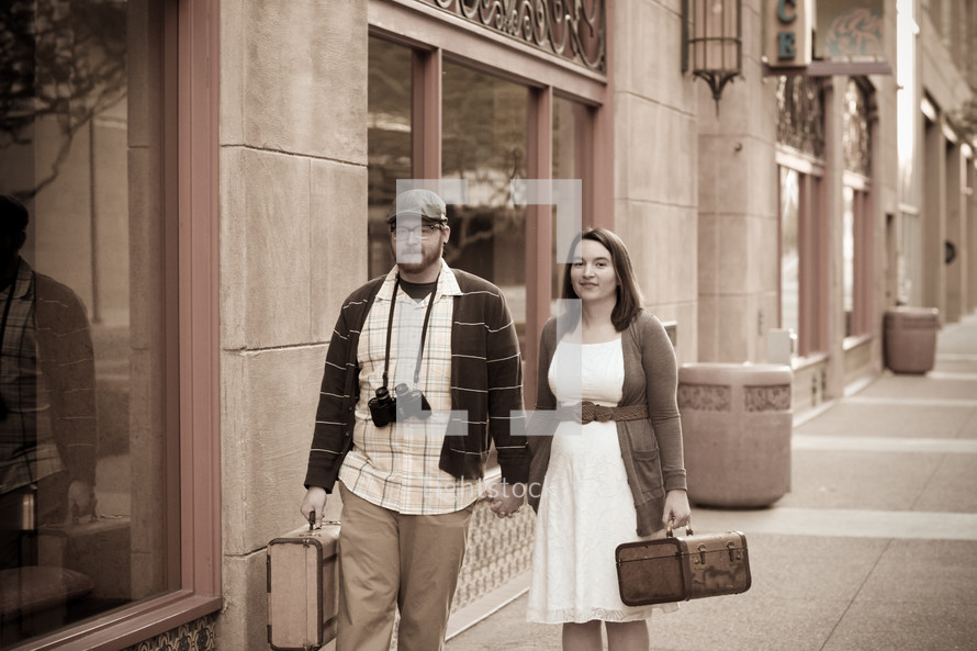 man and a woman holding hands carrying luggage walking down a sidewalk 