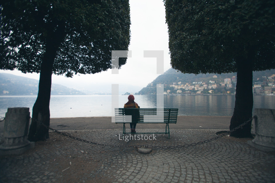 a woman sitting on a park bench looking out at a bay under fog 