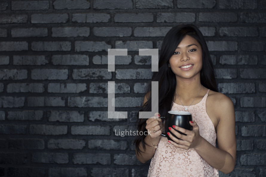 A smiling young woman holding coffee cup and standing against a gray brick wall.