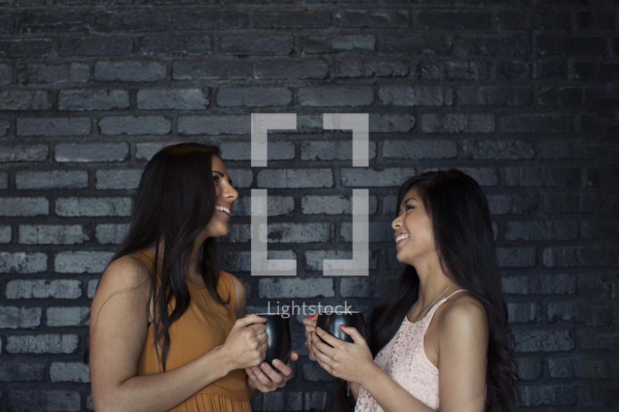 Two young women holding coffee cups and smiling at each other.