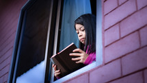 a girl child reading a Bible in an open window during quarantine 