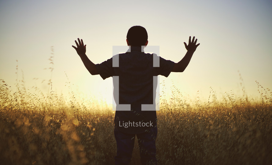 a young man with raised arms walking through a field of tall grass at sunset 