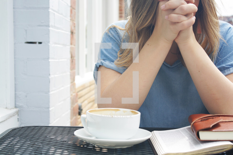 a woman sitting at an outdoor table with a coffee cup, Bible, and journal praying 