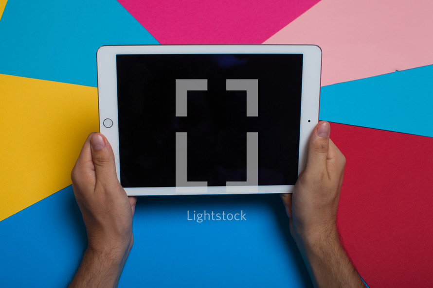 Hands holding an electronic tablet on a multi-colored background.