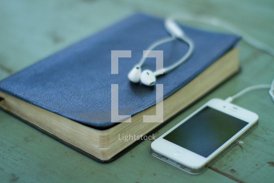 Iphone on a wooden table with earbuds on a closed Bible.