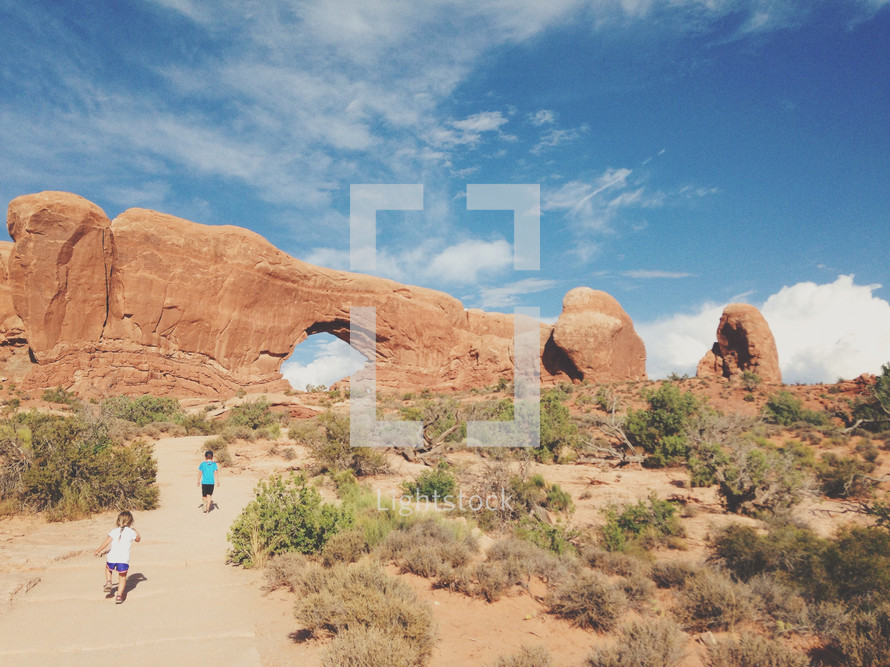 Two children walking a path toward an enormous red rock formation in the desert.