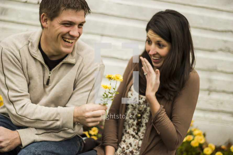 Boy and girl sitting by flowers and a building; he's giving her flowers; she's showing off her engagement ring