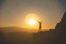 a woman with raised hands standing at the edge of a mountain at sunset 