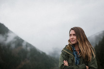 a woman with arms crossed over her chest standing in front of a foggy mountain 