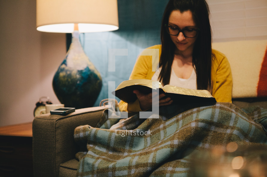 A woman sitting on a couch under a warm blanket reading a Bible. 