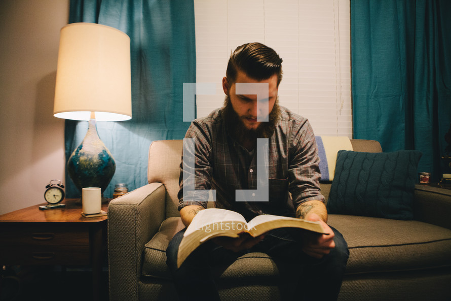 A man reading a Bible while sitting on a couch in his living room. 