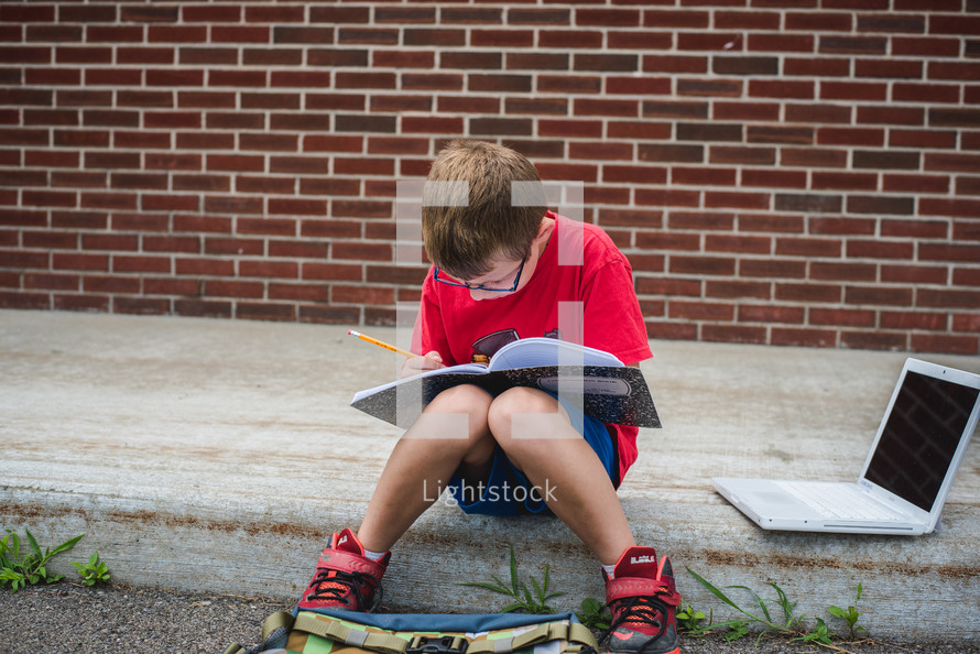 a boy doing his homework sitting on a curb 