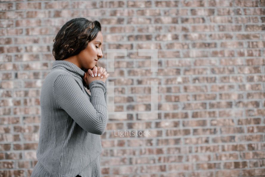 side profile of a woman with praying hands in front of a brick wall 