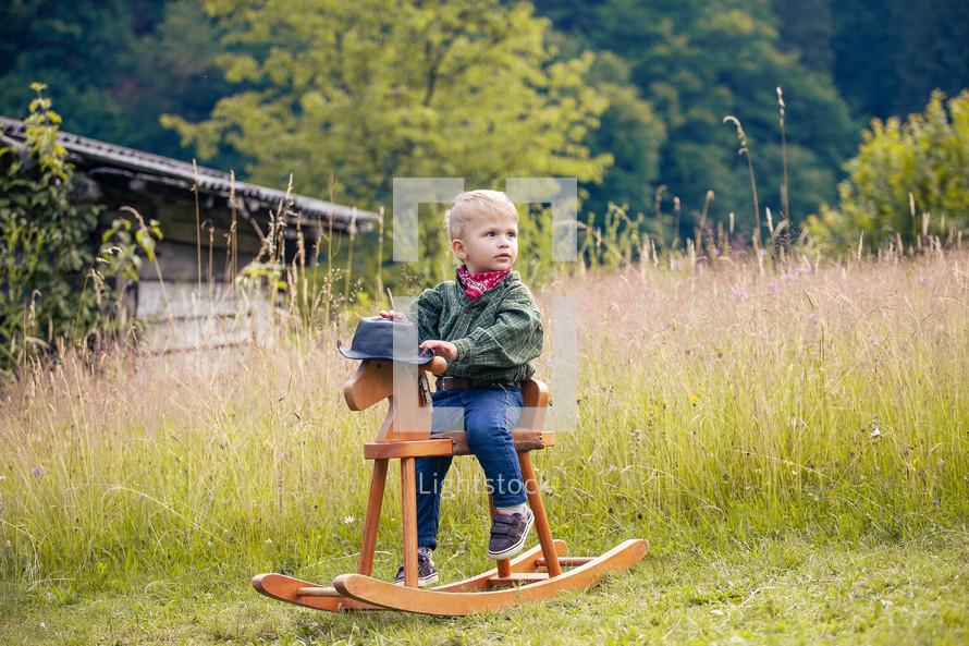 Little Cute Boy in Cowboy Costume on Rocking Horse in the Green Summer Forest