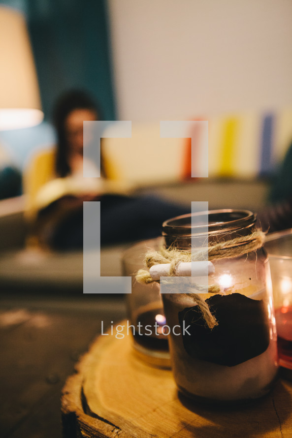 A candle in a mason jar and a woman reading a Bible on a couch in the distance. 