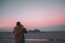 a man standing on a beach at sunset taking pictures 