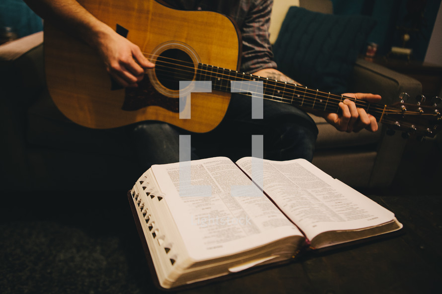 A man sitting on the couch playing his guitar in front of an open Bible. 