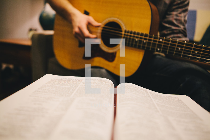 A man playing a guitar while looking at a Bible. 