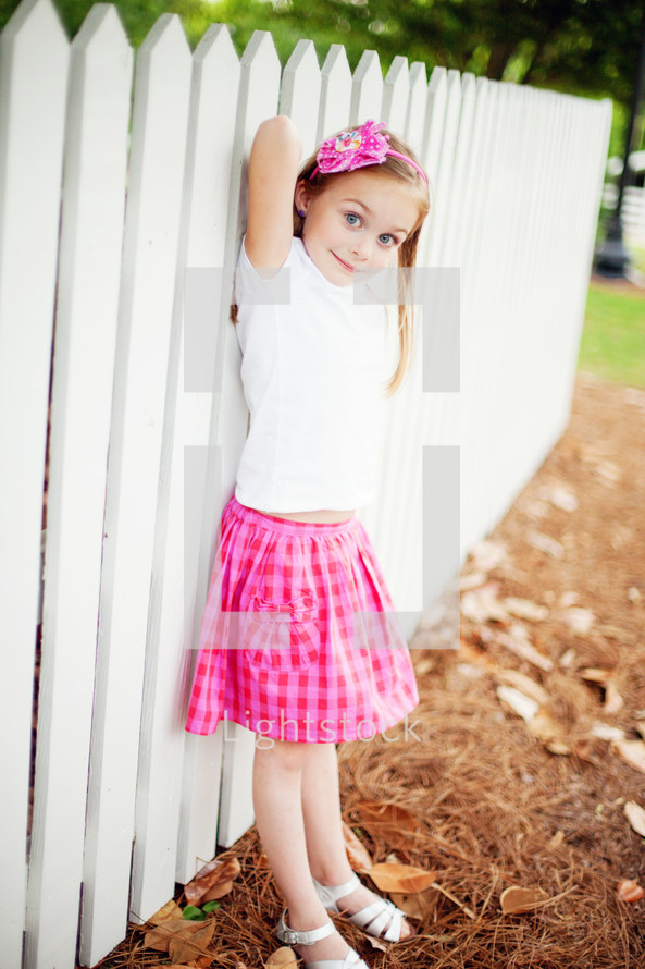 girl child in a gingham skirt standing in front of a white picket fence 