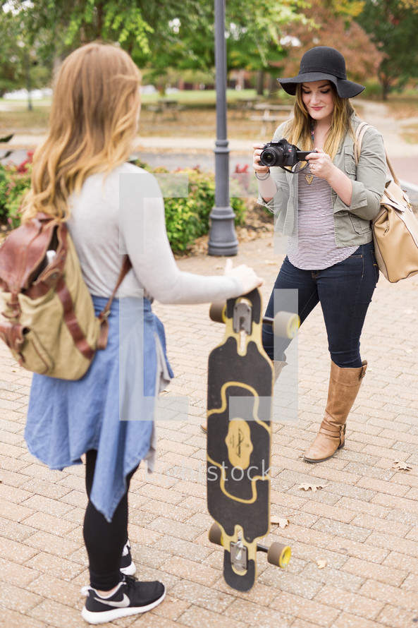 a teen girl holding a skateboard and a friend with a camera 