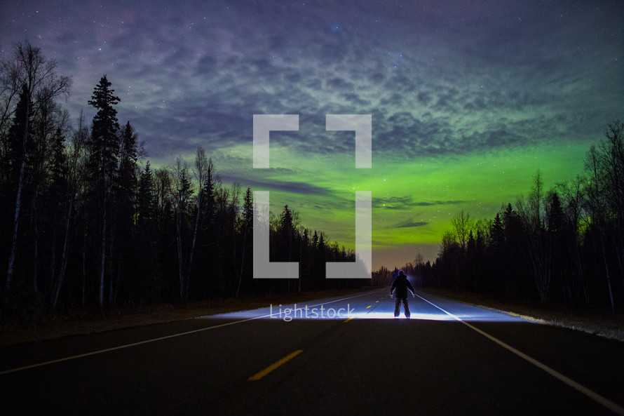 man with a flashlight standing in the middle of a road under an aurora 