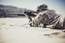 Soldier aiming gun while lying on rock.